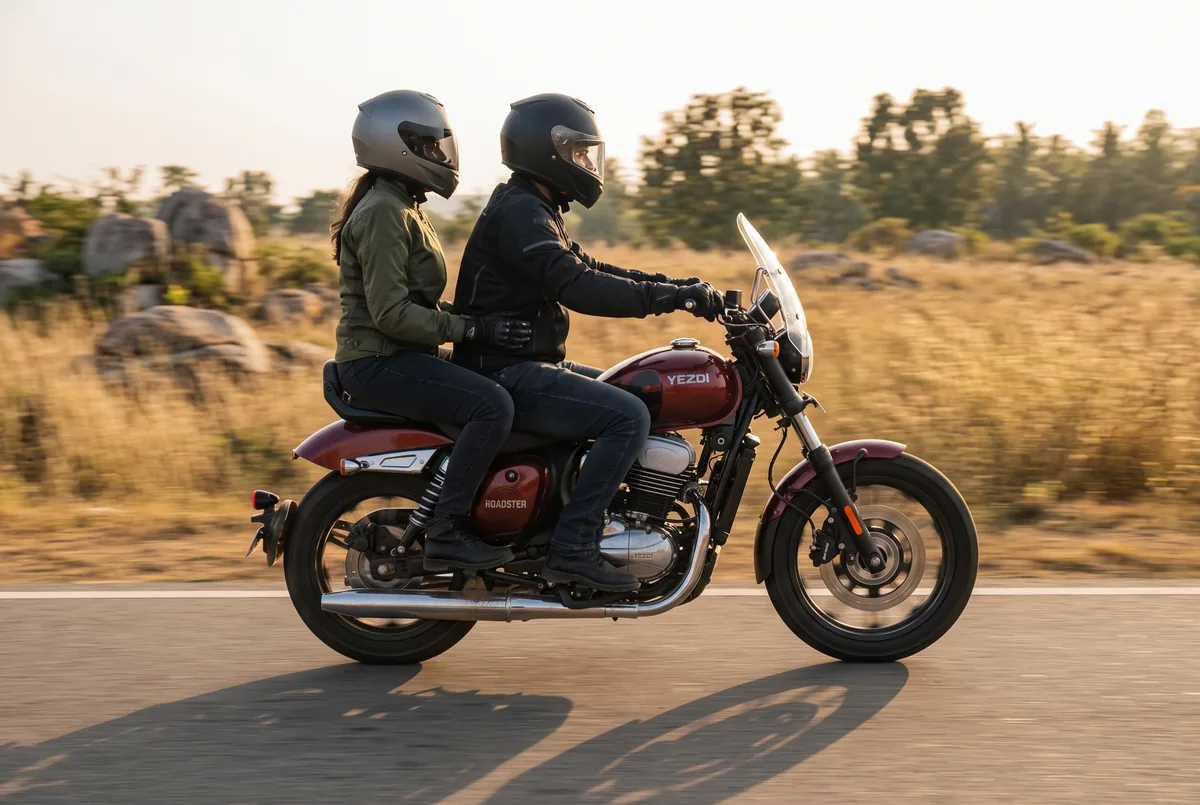 A rider and pillion on a Red Wolf Yezdi Roadster on an open plateau road outside Bengaluru at sunset.