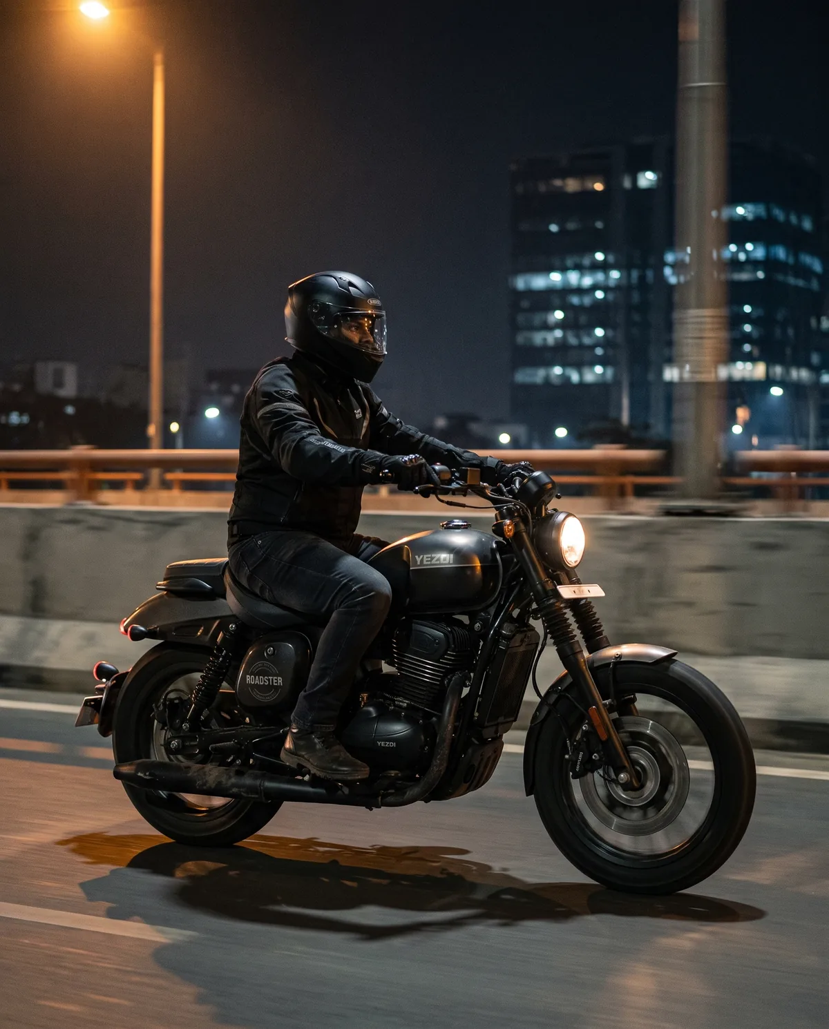 A rider on a Smoke Grey Yezdi Roadster on a Bengaluru flyover at night, towers lit behind.