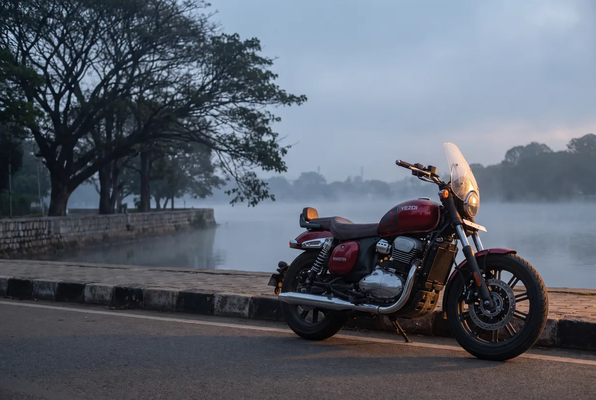 A Red Wolf Yezdi Roadster parked on a lake bund road at dawn, mist over the water.