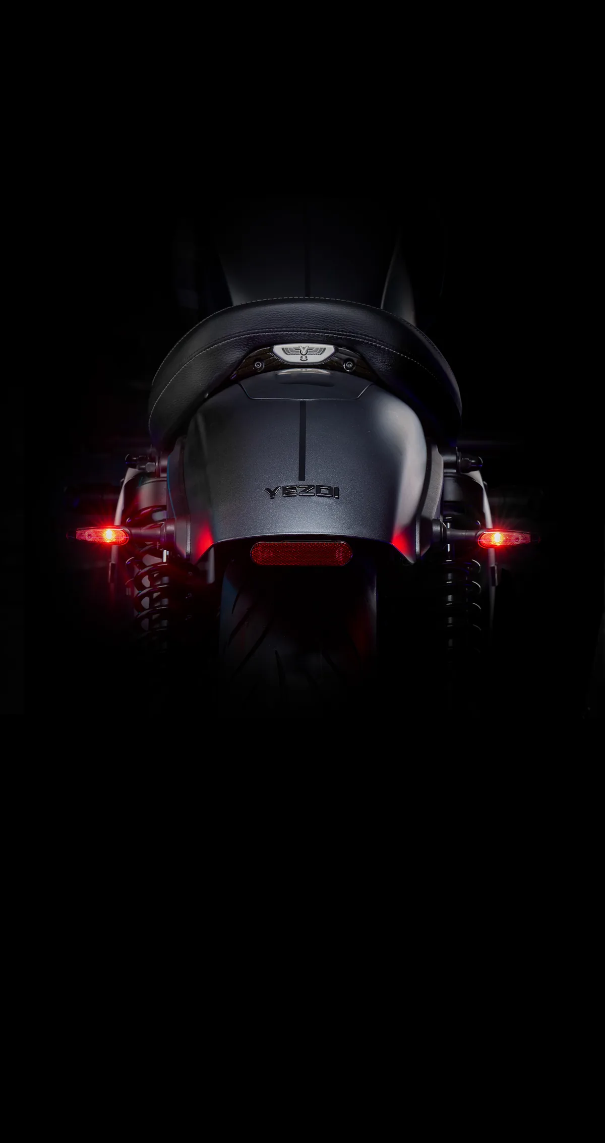 Rear of a matte grey Yezdi Roadster with its tail lamps lit in darkness.